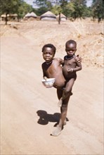 On the Gambaga-Walewale road'. A naked child carrying a bowl balances a toddler on his/her hip on