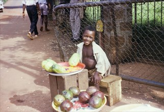 Young fruit seller. A boy sits at a street stall selling fruit to passers-by. Sierra Leone, circa
