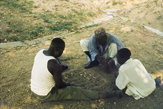 Playing a mancala game, Nigeria. Two men sit on the ground, playing a mancala game in the sand