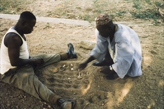 Playing a mancala game, Nigeria. Two men sit on the ground, playing a mancala game in the sand.