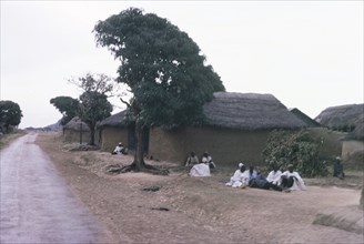 A Hausa village. Inhabitants of a Hausa village sit on the ground outside several square,