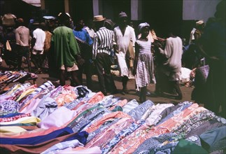 Cloth for sale in Freetown. Shoppers walk past a colourful market stall selling patterned rolls of