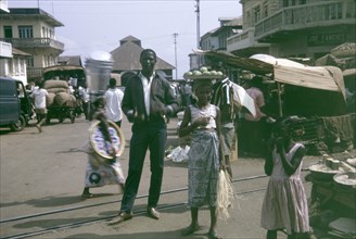 Market scene at Freetown, Sierra Leone. A woman balances a platter of fruit on her head, as she