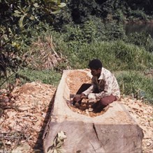 Carving out a canoe. A carpenter sits on top of a large piece of timber that he is carving out to