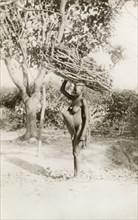 Fuel gatherer'. Profile shot of a semi-naked African woman. She stands, balancing a large bundle of