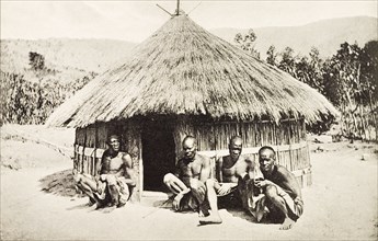 Vernacular hut in Kenya. Four men squat on the ground in front of a round hut with a thatched roof.