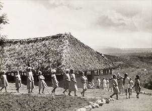 Kikuyu schoolchildren. A group of Kikuyu schoolchildren walk in single file towards a thatched roof
