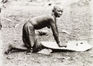 Grinding millet. A Kikuyu woman, naked from the waist up, kneels on the ground as she grinds millet