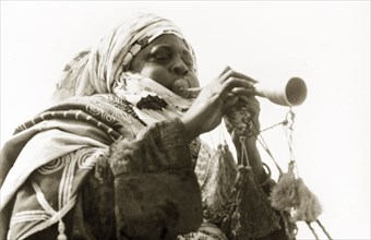 A Nigerian musician. A Nigerian musician plays a trumpet decorated with tassels. Nigeria, circa