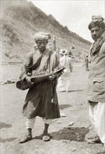 Playing a sarod at the border. An Indian man, armed with a rifle and wearing a bandolier, plucks a