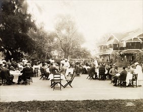 A formal garden party. Small groups of European men and women sit around dining tables on a tennis