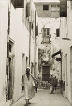 A back street of Lamu. A man in traditional Arab dress, including a kurta (shirt) and a fez, walks