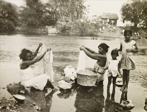 Washing clothes in a stream. Two female plantation workers, accompanied by two young girls, sit in
