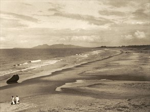 Malabar Coast and Mount Deli. Two tiny figures walk along a vast sandy beach on the Malabar Coast