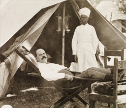 Taking a break at a camp site, India. James Murray, attended to by an Indian servant, reclines in a