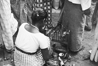 Short-eats' in Ceylon. A female market trader squats at her stall, apparently preparing some sort