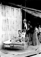 Baby in a wooden cradle, Burma. A Burmese lady amuses a baby with a toy as it lies in a hanging