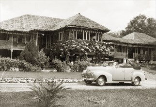 A stilted bungalow in British Malaya. A small car is parked in the driveway outside Dr Tweedie's