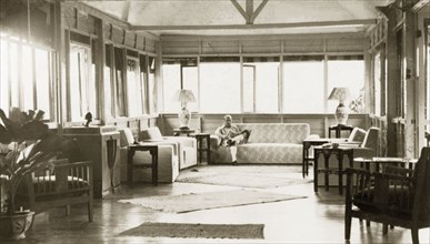 Drawing room of a colonial bungalow, British Malaya. Dr Reid Tweedie sits reading a newspaper in
