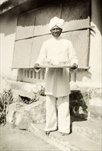 An Indian house servant. An Indian house servant poses on the steps of a colonial bungalow holding
