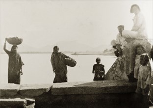 Women at a lakeside ghat, India. Women carry loaded baskets up a ghat (stepped wharf) after washing