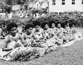 Women perform a 'vakamalolo'. Ten young Fijian women sit cross-legged in a line and clasp their