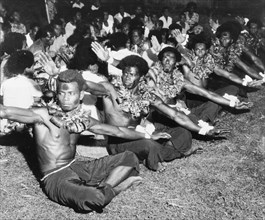 Men perform a 'vakamalolo'. A group of young Fijian men sit cross-legged in a line and stretch out