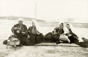 Women in traditional Palestinian attire. Four women sit on mats on a rooftop as they drink coffee