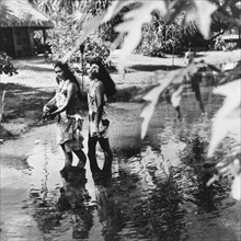 Two Tahitian women. Two Tahitian women walk along a shallow stream, wearing floral print 'pareus'