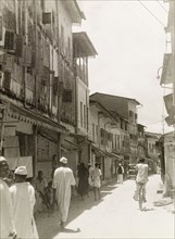Commercial street in Stone Town. Tall, narrow shops with shuttered windows and awnings flank a