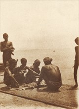 Boatmen on the breakwater. Boatmen cut up fish on the breakwater. Mormugao, Goa, India, circa 1906.