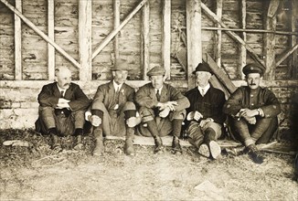 Smoking break at Hardwick Estate. Five men from a shooting party sit against a wall, smoking,
