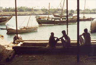 Dhows in Kilindini harbour'. Three men sit in the shade at Kilindini harbour, watching the dhows as