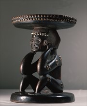 Caryatid stool supported by a depiction of a matrilineal ancestor, her seated posture with hands behind the ears indicates mourning, reminding the living of the necessity of sacrifices to the dead