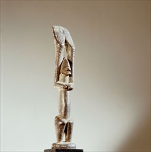 A wood sculpture of a nommo figure with hands up raised, the offspring of the Dogon creator god, Amma and his Earth wife