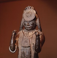 A large figure, one of the Tsoede bronzes, wearing an outer garment covered with cowrie shells, a necklace of leopards teeth and a pectoral with representations of a ram and birds
