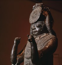A large figure, one of the Tsoede bronzes, wearing an outer garment covered with cowrie shells, a necklace of leopards teeth and a pectoral with representations of a ram and birds