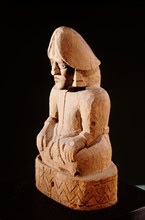 Seated wooden figure with large earrings and a phallus shaped headdress