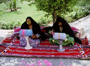 Two women wearing black veils and burqa face masks prepare coloured braids