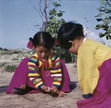 Two young Korean girls in brightly coloured national dress, crouching on the ground as they play a game with small stones