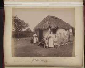 Women and children posed outside thatched hut