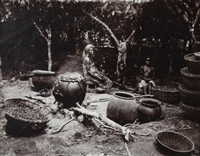 View of seated figures with baskets and clay pots, one cooking palm kernels, in cultivated grove