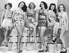 United States:  c. 1952.
Six attractive young women in two piece bathing suits sit in a row on