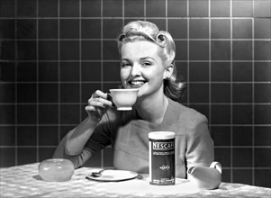 United States:  c. 1960.
A single woman at a kitchen table enjoys a cup of Nescafe.