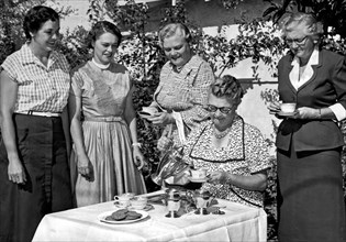 California:   c. 1948.
Four women gather around a table outdoors while another one serves coffee