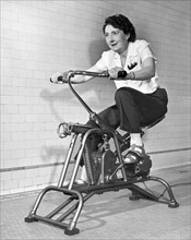 United States: c. 1960.
An older woman pedals on her exercycle.