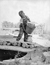 A small boy on a farm pours water from a bucket into a pitcher for the house.