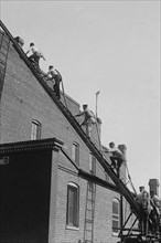A team of Firefighters with Hoses on Their backs climbs a ladder 1920