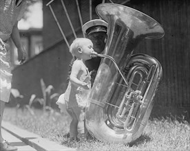 An nearly naked infant toots a tuba many times his size 1917