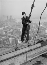 Cameraman in suit holds onto cable as he walks unharnessed over a skyscraper's steel girders